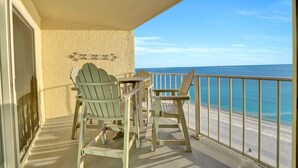 Ein Balkon mit zwei Adirondack-Stühlen und einem kleinen Tisch, mit Blick auf einen Strand und klares, blaues Wasser.