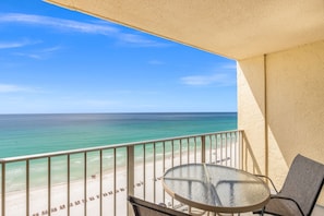 A balcony with a glass table and chairs, overlooking a beach with a clear view of the ocean.