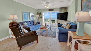 Een woonkamer met twee banken, een salontafel en een televisie. Er is een balkon met uitzicht op de oceaan.