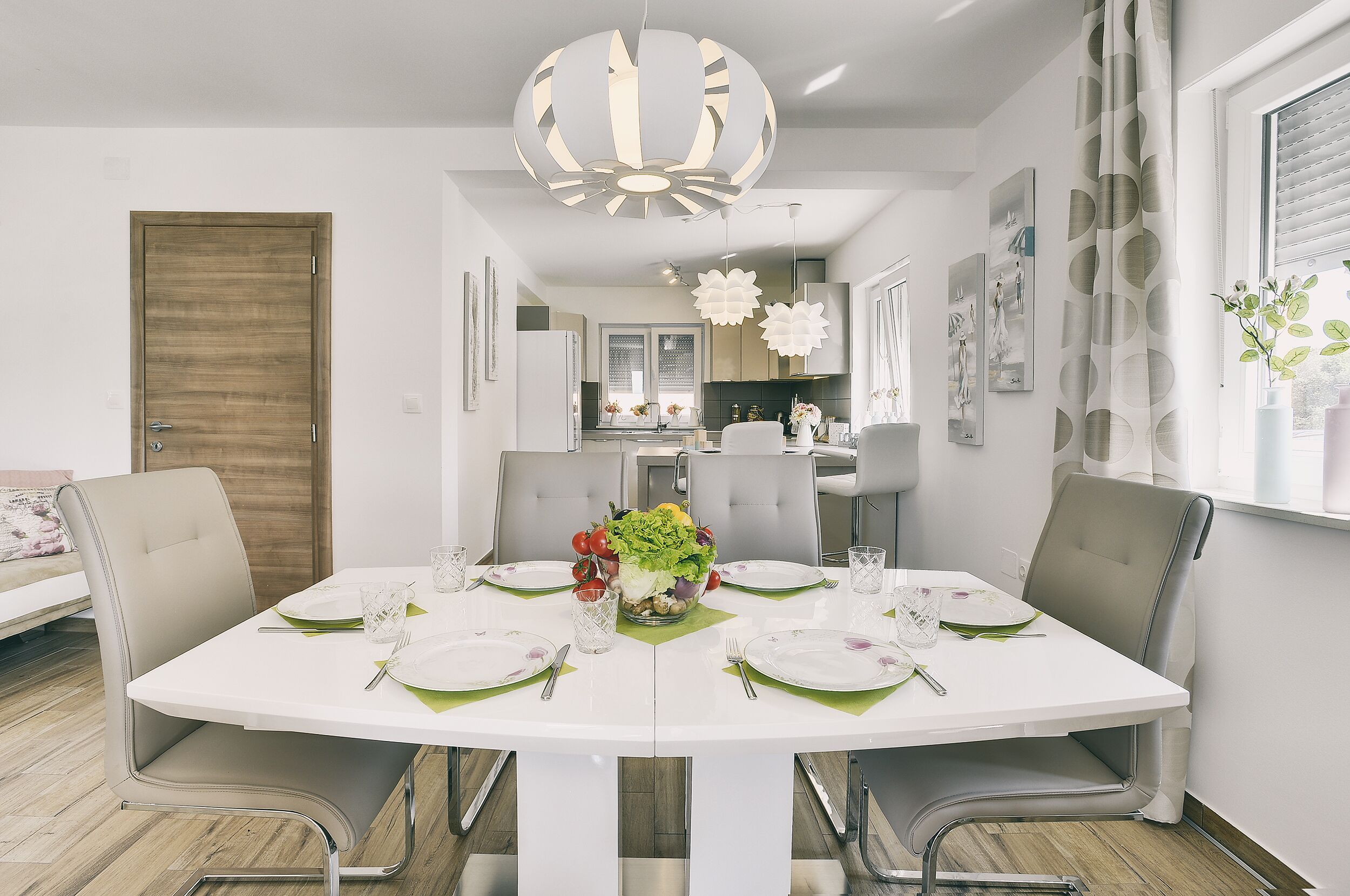 Uma sala de jantar com uma mesa branca posta para quatro pessoas, rodeada por cadeiras modernas, uma luminária decorativa no teto e vista para a cozinha.