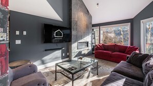 Ett modernt vardagsrum med en platt-TV, ett soffbord i glas, en röd soffa och ett stort fönster med utsikt över träd.