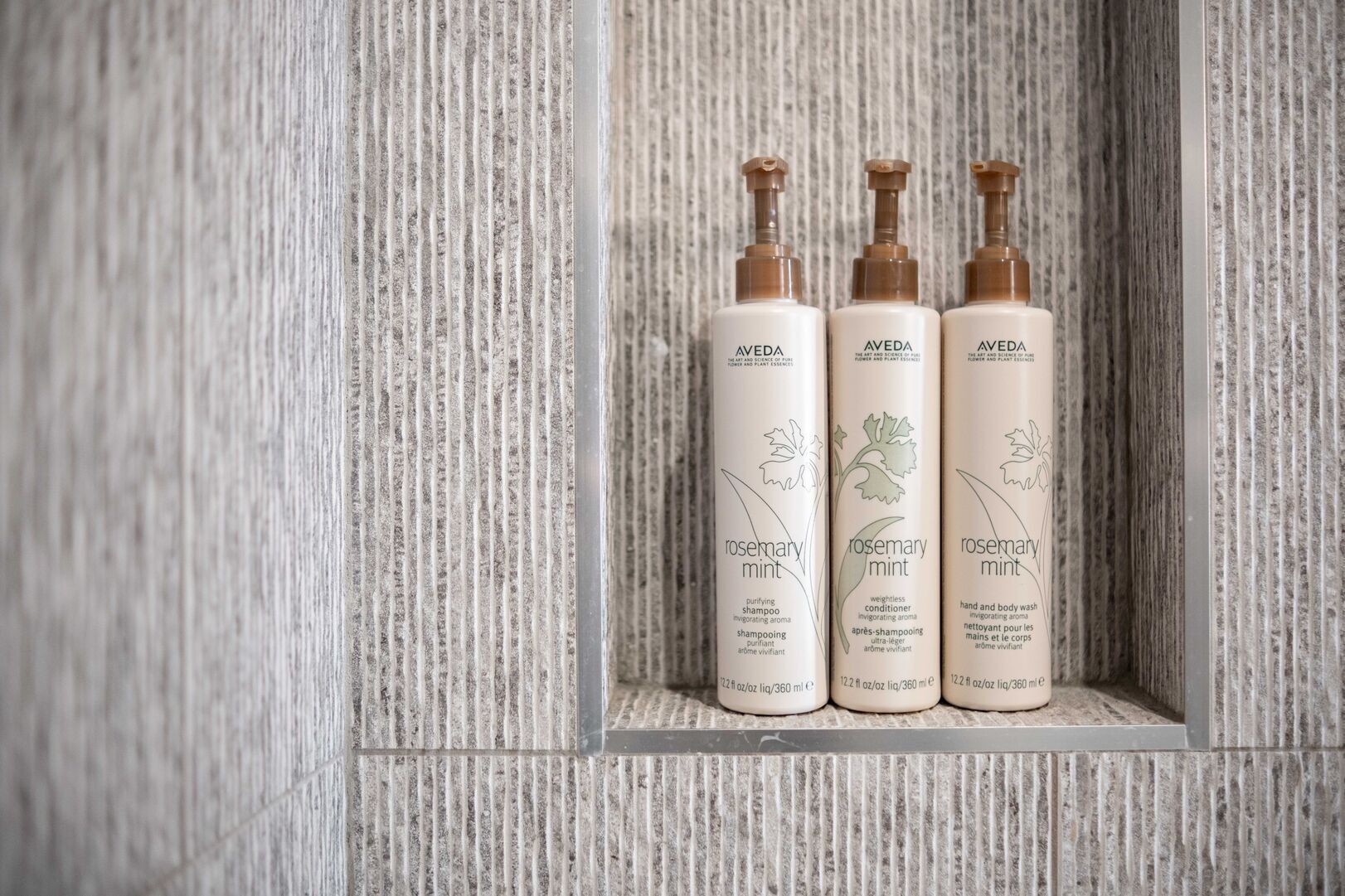 Drei Flaschen Aveda Rosmarin-Minze-Shampoo und -Spülung auf einem strukturierten, hellen Fliesenregal.