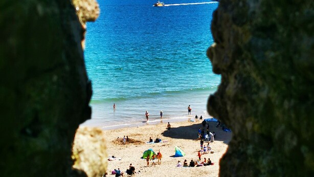 A beach scene viewed through a natural rock opening, with clear blue water, a sandy shore, and a few people and umbrellas. - Stylish Family-Friendly Sea-View Home, sleeps 9 (Tenby)