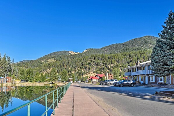 Un hotel frente al lago, con su reflejo en el agua, una carretera con coches aparcados y montañas al fondo.