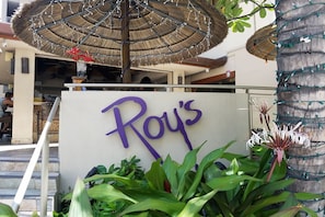 Roy's features a charming outdoor seating area with lush greenery and a thatched umbrella.