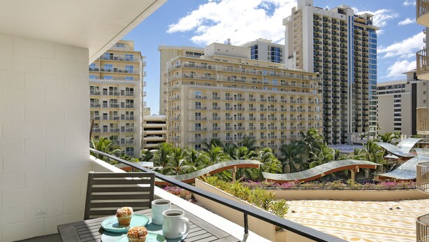 A balcony with a table set for two, overlooking a resort area with palm trees and a pool. - Modern condo in Heart of Waikiki! 1 block to beach, AC, Wi-Fi, free parking! (Honolulu)