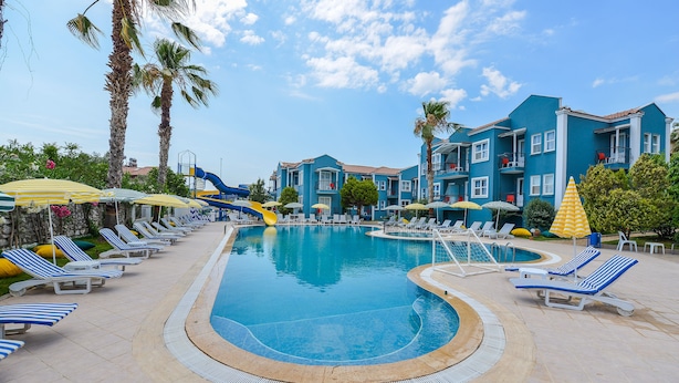 A swimming pool area with lounge chairs and umbrellas, surrounded by palm trees and a building with balconies. - BLUE PEARL HOTEL& VILLAS (Fethiye)