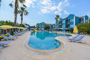 Area piscina con sedie a sdraio e ombrelloni, circondata da palme e un edificio con balconi.