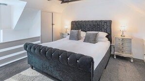 Un dormitorio con cama y cabecera capitonada, dos mesitas de noche con lámparas, una puerta blanca y techo inclinado con vigas a la vista.