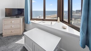 A room with a large bay window overlooking a beach, a television on a wicker dresser, and a white radiator. - 9400 Building - Oceanfront Condo with Panoramic views of the ocean and bay! (Ocean City)