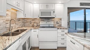 A modern kitchen with white cabinets, a stainless steel sink, a built-in microwave, and a stove. There's a view of the ocean through a balcony. - 9400 Building - Oceanfront Condo with Panoramic views of the ocean and bay! (Ocean City)