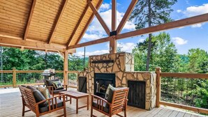 A covered deck with wooden furniture, a stone fireplace, and a view of the surrounding forest. - Red Cedar Lodge #396 (Sevierville)