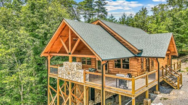 A wooden cabin with a stone foundation, a covered porch, and a steeply pitched roof, surrounded by dense forest. - Red Cedar Lodge #396 (Sevierville)