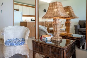 A wicker chair with a blue cushion, a wicker lamp on a glass table, and a stack of books. - Leinaala Oceanfront #303 Panoramic Ocean Front View, A/C Great Location, Pool (Kihei)