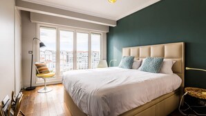 Ein Schlafzimmer mit einem großen Bett, Blick auf die Stadt durch große Fenster, einer Stehlampe und einem Sessel.