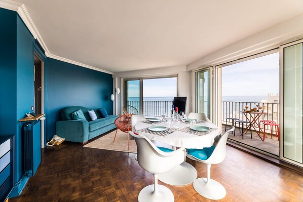 Uma sala de estar moderna com uma parede azul de destaque, um sofá e um espaço de jantar com uma mesa posta para quatro pessoas.
