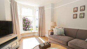 A living room with a TV, sofa, coffee table, and a window with curtains. - Snowdrop Cottage (Windermere)