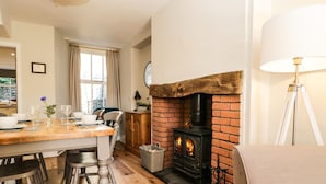 A cozy living room with a brick fireplace, wooden flooring, and a dining area with chairs and a table set for two. - Snowdrop Cottage (Windermere)