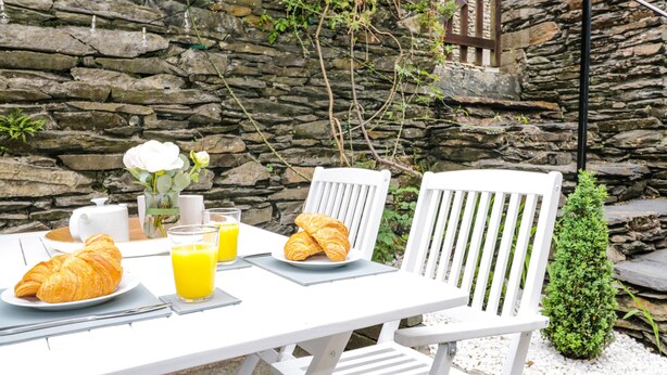 A stone wall, white chairs, a table with croissants, juice, and a vase with flowers. - Snowdrop Cottage (Windermere)