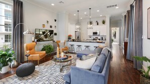 A modern living room with a sofa, armchair, and ottoman, a kitchen in the background, and a dining area with bar stools.