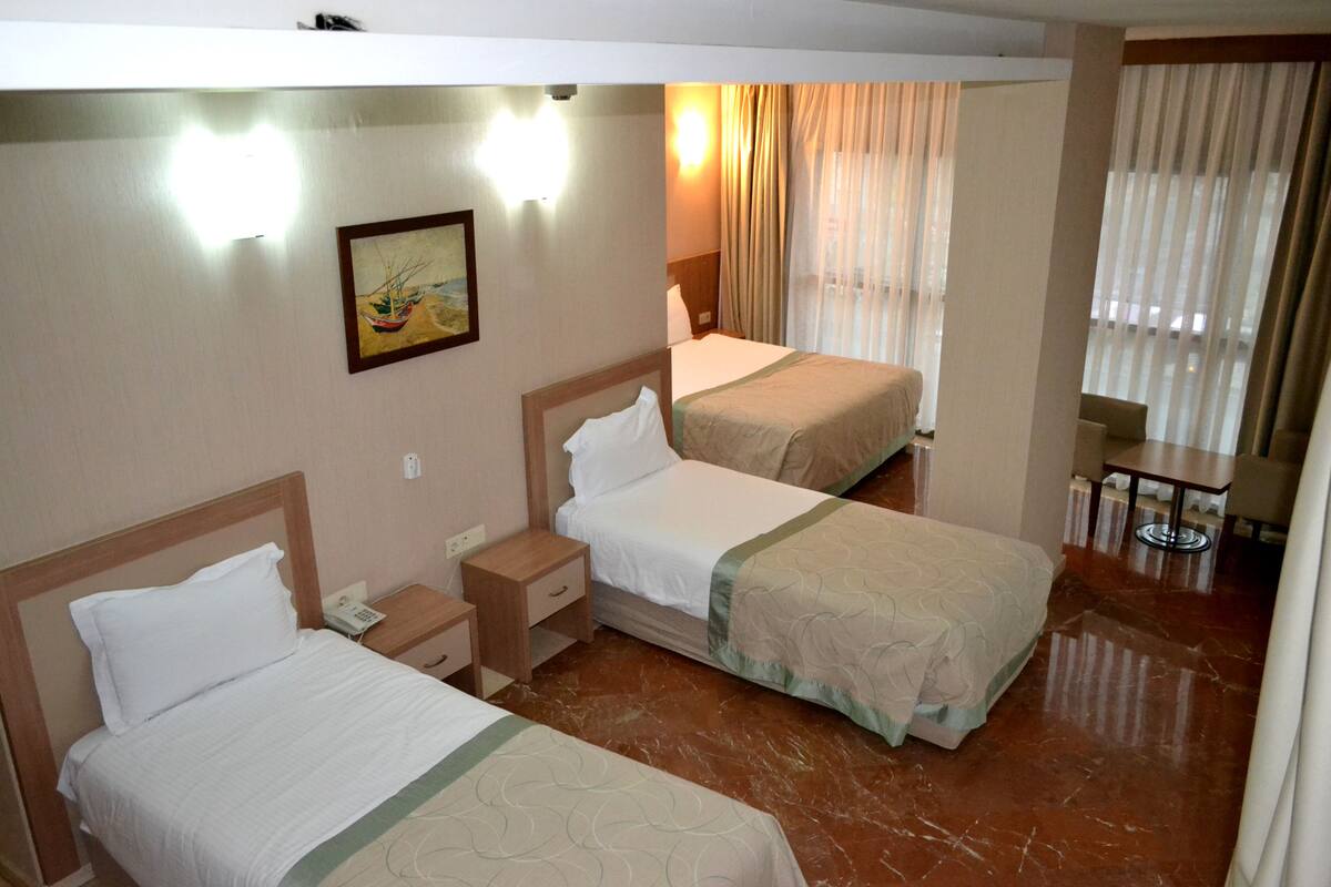A hotel room with two beds, a bedside table with a phone, a painting on the wall, and a window with curtains.