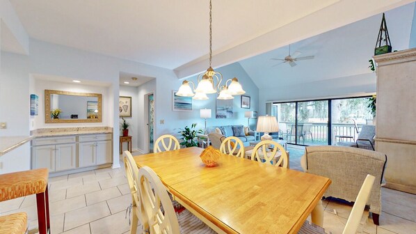 A spacious living area with a wooden dining table, white chairs, and a ceiling fan.