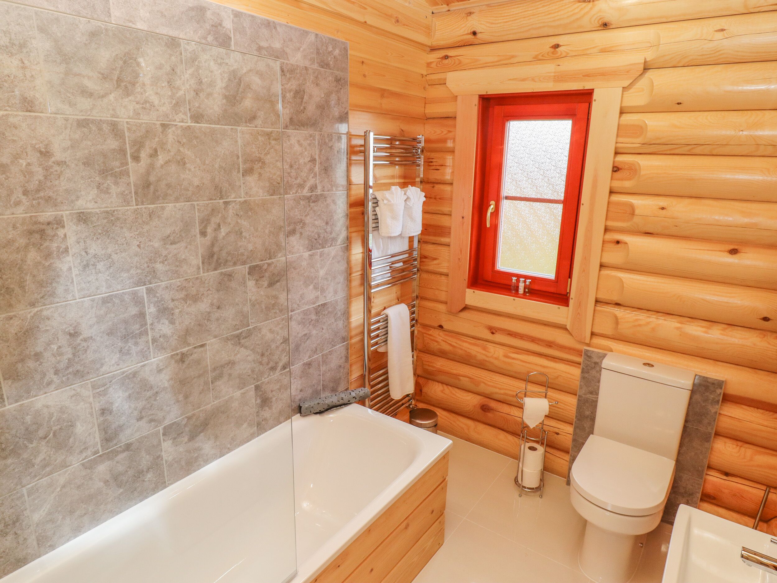 A wooden cabin bathroom with a bathtub, toilet, and a window with a red frame.