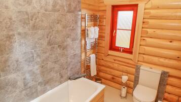 A wooden cabin bathroom with a bathtub, toilet, and a window with a red frame.