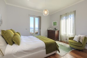 A bedroom with a large bed, a green armchair, a dresser, and a view of the city through the window. - Villa Ramella (Baveno)