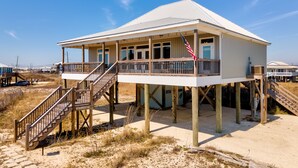 Una casa frente al mar con balcón, escaleras de madera y una playa arenosa al fondo.