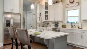 Una cocina moderna con armarios blancos, un refrigerador de acero inoxidable, una isla central con taburetes de bar, iluminación colgante y un reloj en la pared.