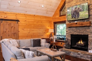 A cozy cabin interior with a stone fireplace, wooden walls, a large TV mounted above it, and a comfortable seating area. - Riverside Relaxation, Fireside Moments & Serene Sips in Mountain Breezes - Toccoa River Mist (Blue Ridge)
