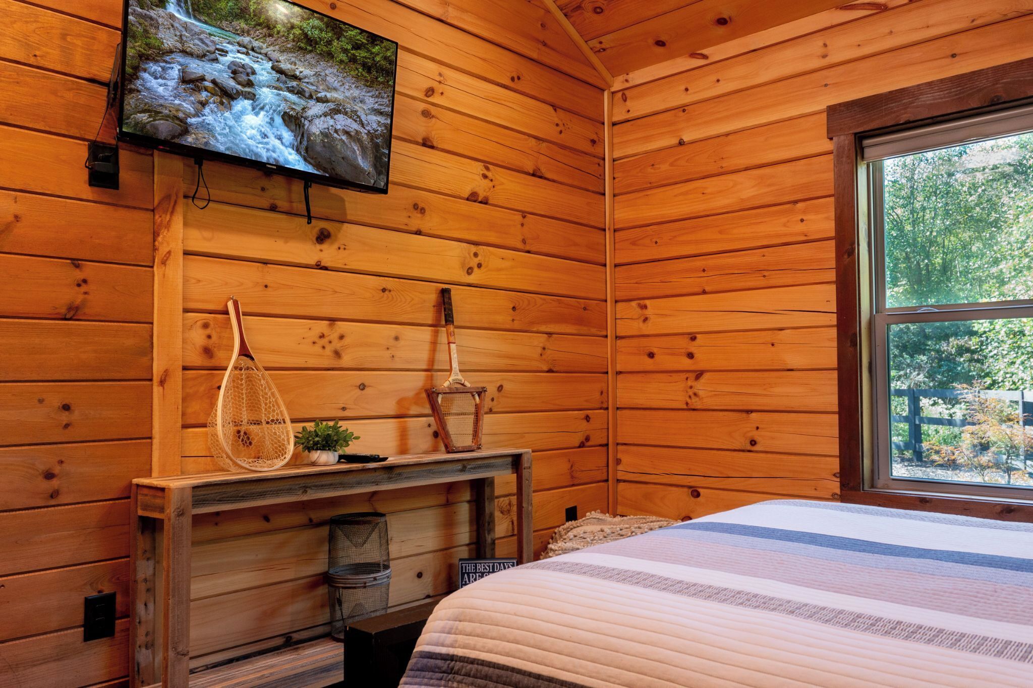 Habitación de cabaña de madera con cama, televisor montado, una mesita con una cesta y ventana con vista a árboles.
