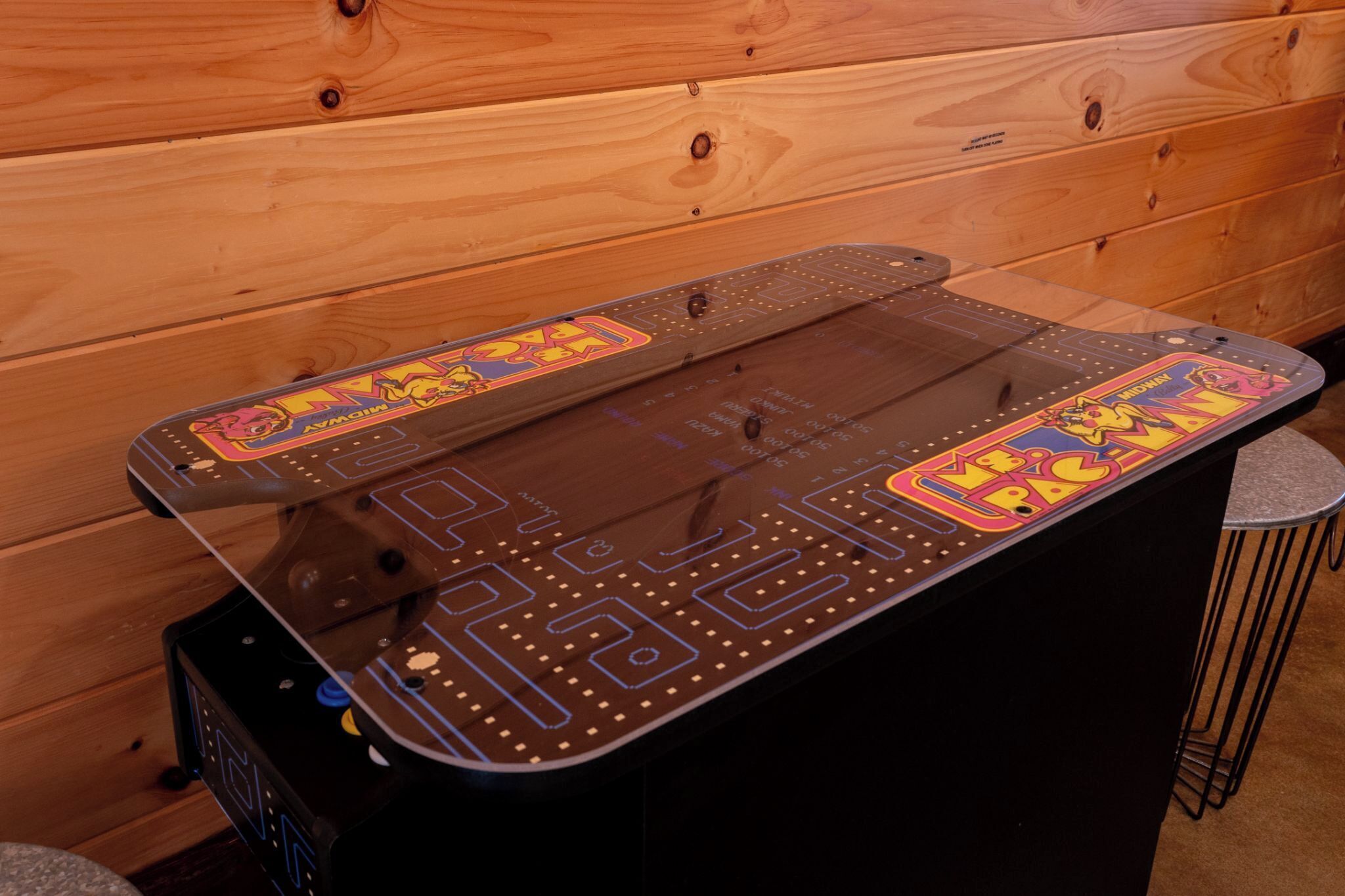 A pool table with a colorful design, a wooden wall in the background, and a metal bar stool to the right.
