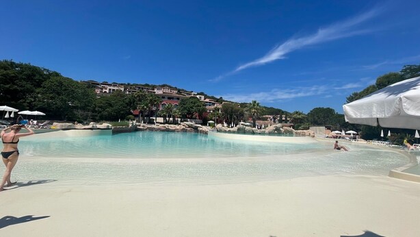 Ein Poolbereich mit klarem Wasser, umgeben von einem Sandstrand und mit Gebäuden und Bäumen im Hintergrund.