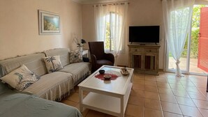 Ein Wohnzimmer mit Sofa, Sesseln, Couchtisch, Fernseher und einer Schiebetür zum Außenbereich.