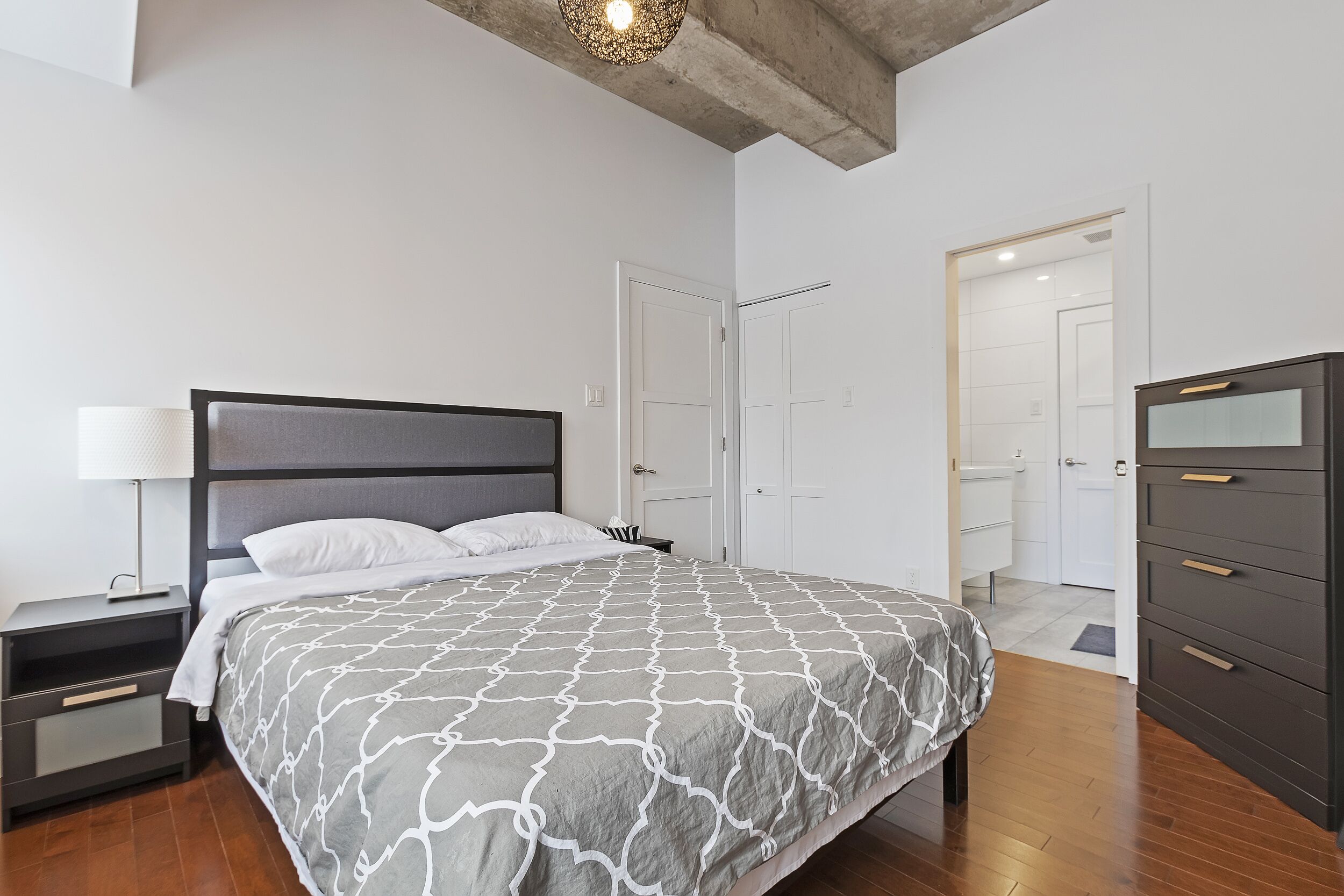 A modern bedroom with a bed, nightstand, dresser, and a door leading to a bathroom.