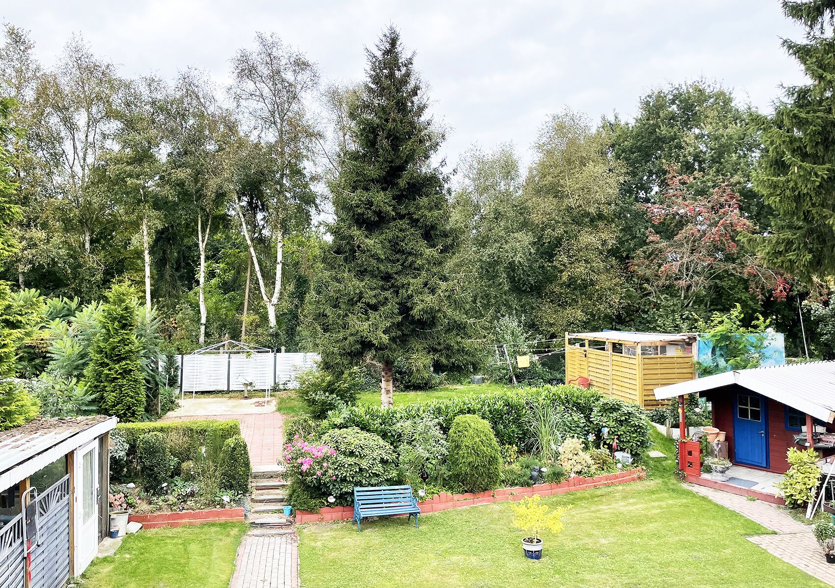Een verzorgde tuin met een pad dat leidt naar een houten constructie, omringd door verschillende bomen en struiken.