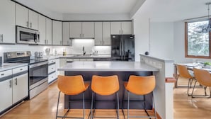 A modern kitchen with white cabinets, stainless steel appliances, and a central island with three orange bar stools. - 5 BR Wintergreen House w/ Game Room, 75” TV, Ping Pong, Foosball, Wet Bar & Deck (Nellysford)