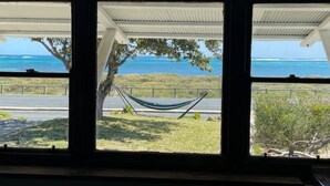 Une vue à travers une fenêtre, avec un hamac, un arbre et une route menant à l’océan.