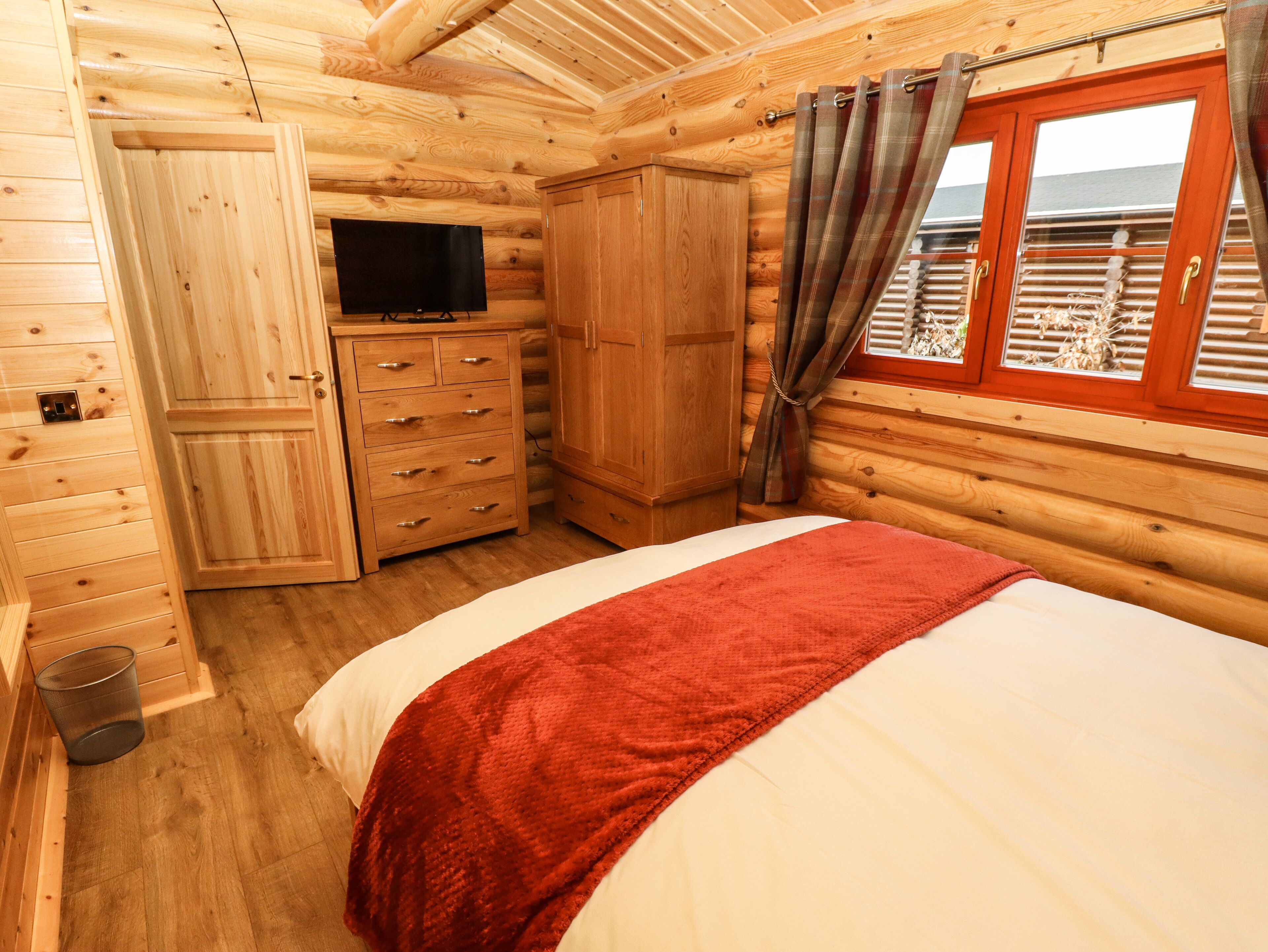 A wooden cabin room with a bed, a TV, a wardrobe, and a window with curtains.