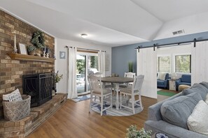 Een woonkamer met een open haard, een eettafel met stoelen, een bank en een groot raam met zonwering.