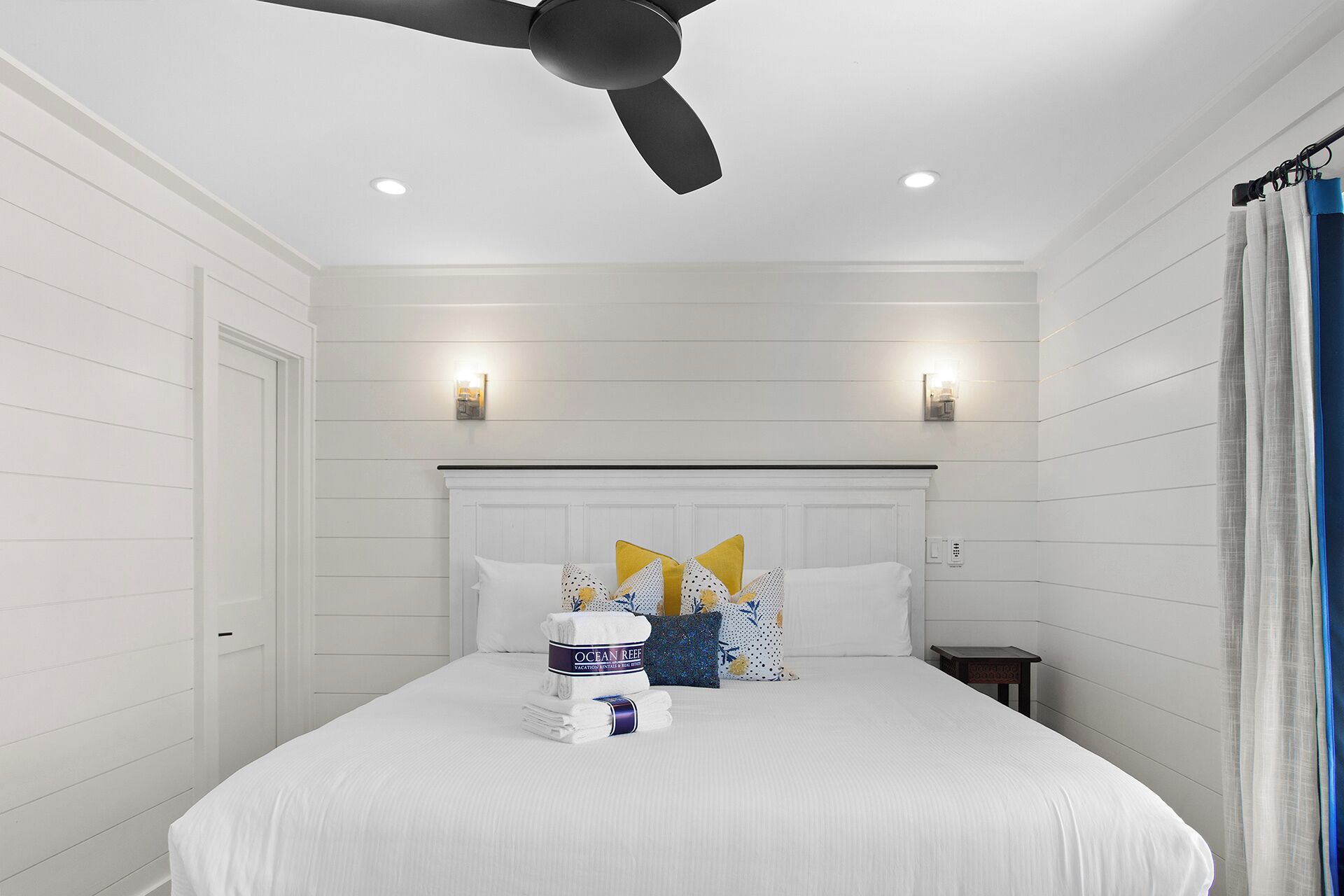 Une chambre avec un lit blanc, un ventilateur de plafond, des spots encastrés et une table de chevet.