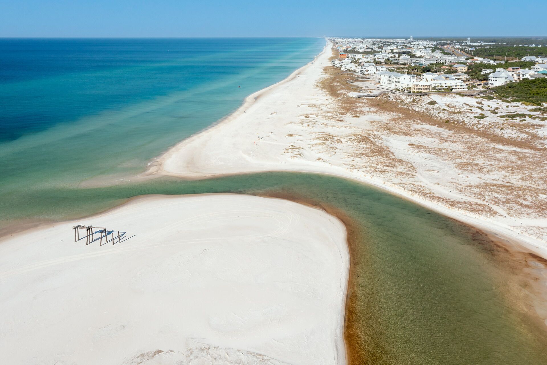 Vista aérea de una zona costera con playa de arena, aguas azules cristalinas y un pueblo con edificios blancos.