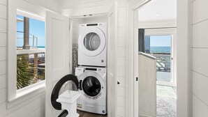 Une buanderie équipée de deux lave-linge et deux sèche-linge Whirlpool, une fenêtre donnant sur la plage et une porte menant à un balcon avec vue sur l’océan.