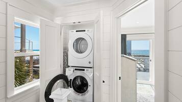 Une buanderie équipée de deux lave-linge et deux sèche-linge Whirlpool, une fenêtre donnant sur la plage et une porte menant à un balcon avec vue sur l’océan.