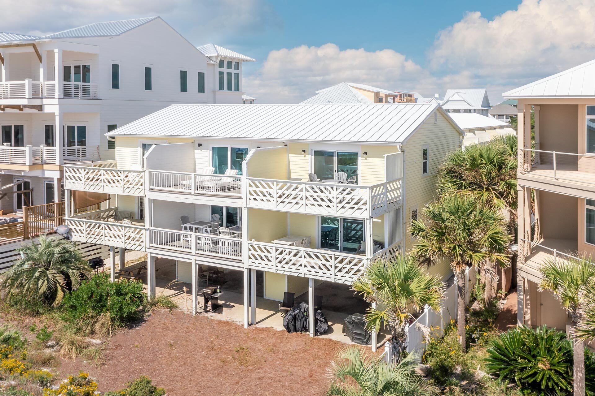 A two-story beachfront house with a balcony, surrounded by palm trees and other vegetation.