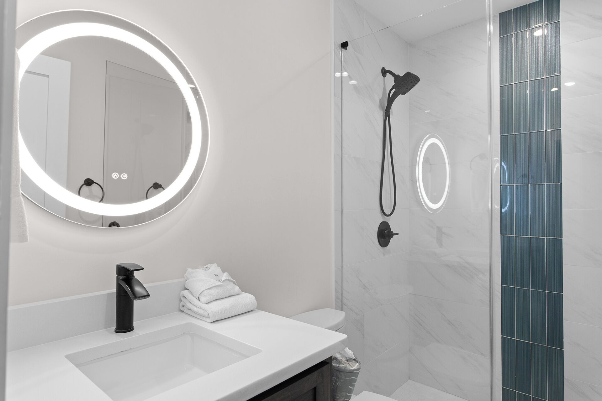 A modern bathroom with a white sink, a round mirror with lights, a shower with a handheld showerhead, and a glass-enclosed shower area.