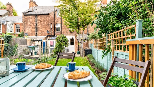 Une table de jardin avec des assiettes remplies de nourriture et des tasses, entourée de verdure et avec un bâtiment en briques en arrière-plan.
