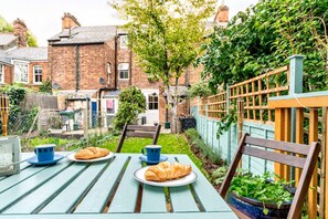 Una mesa en el jardín con platos de comida y vasos, rodeada de follaje y un edificio de ladrillo al fondo.